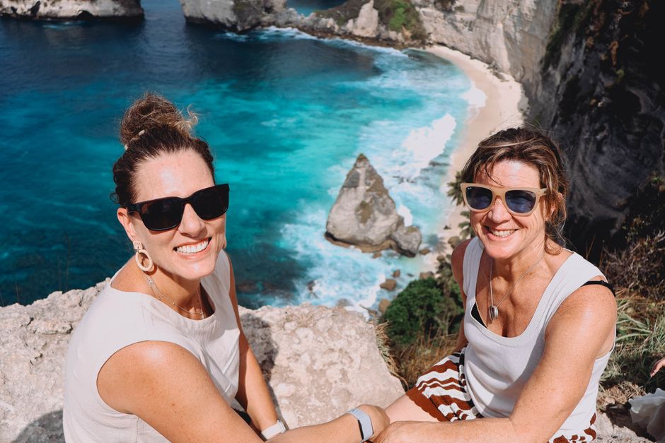 Two women pose happily on a cliffside overlooking Nusa Penida's stunning blue waters in Bali, Indonesia.