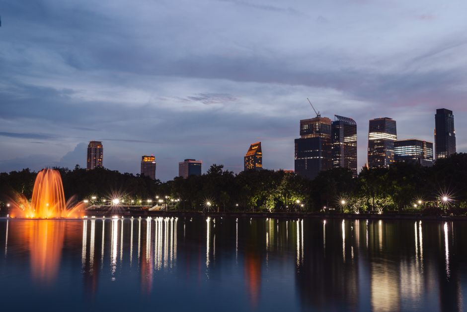 Urban skyline at twilight with a glowing fountain reflecting on a serene lake surface.