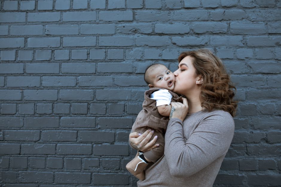 A mother lovingly kisses her baby against a gray brick wall backdrop.
