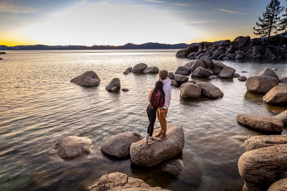Young couple stands on rocks by a peaceful lake at sunset, embracing tranquility.