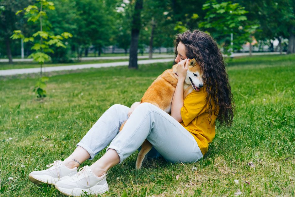 A woman sitting on grass hugging her Shiba Inu dog in a lush green park.