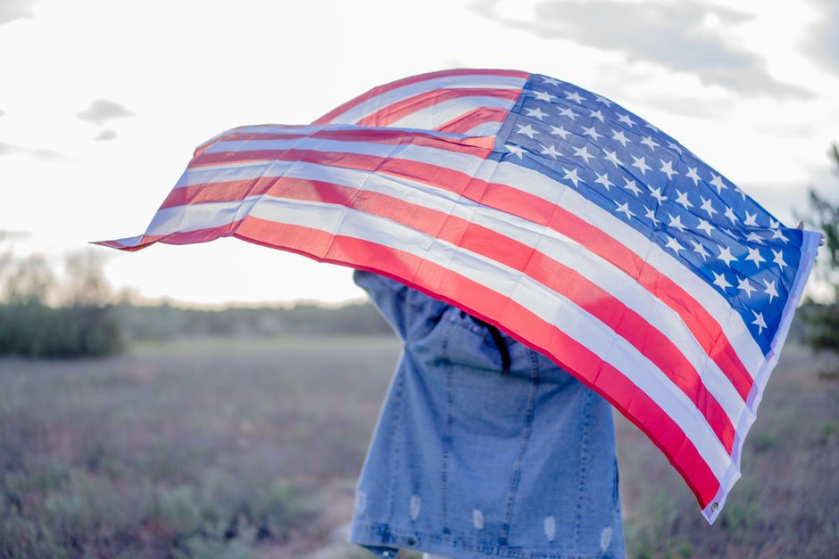 Back view of unrecognizable person in denim jacket with USA flag standing on grassy field in countryside in daytime
