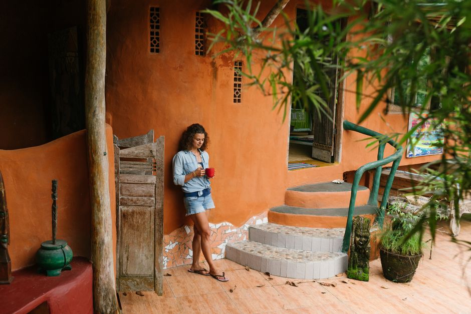 Full body brunette wearing denim clothes with cup of hot drink leaning on rural cottage wall while visiting exotic tropical country